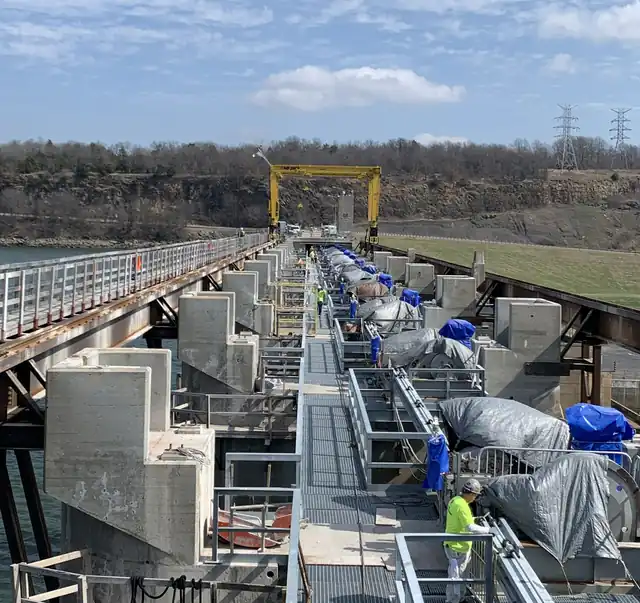 Eufaula dam spillway deck with gate hoists tarped, a gantry crane staged on the walkway, crews in hi-vis, lake and bluff behind
